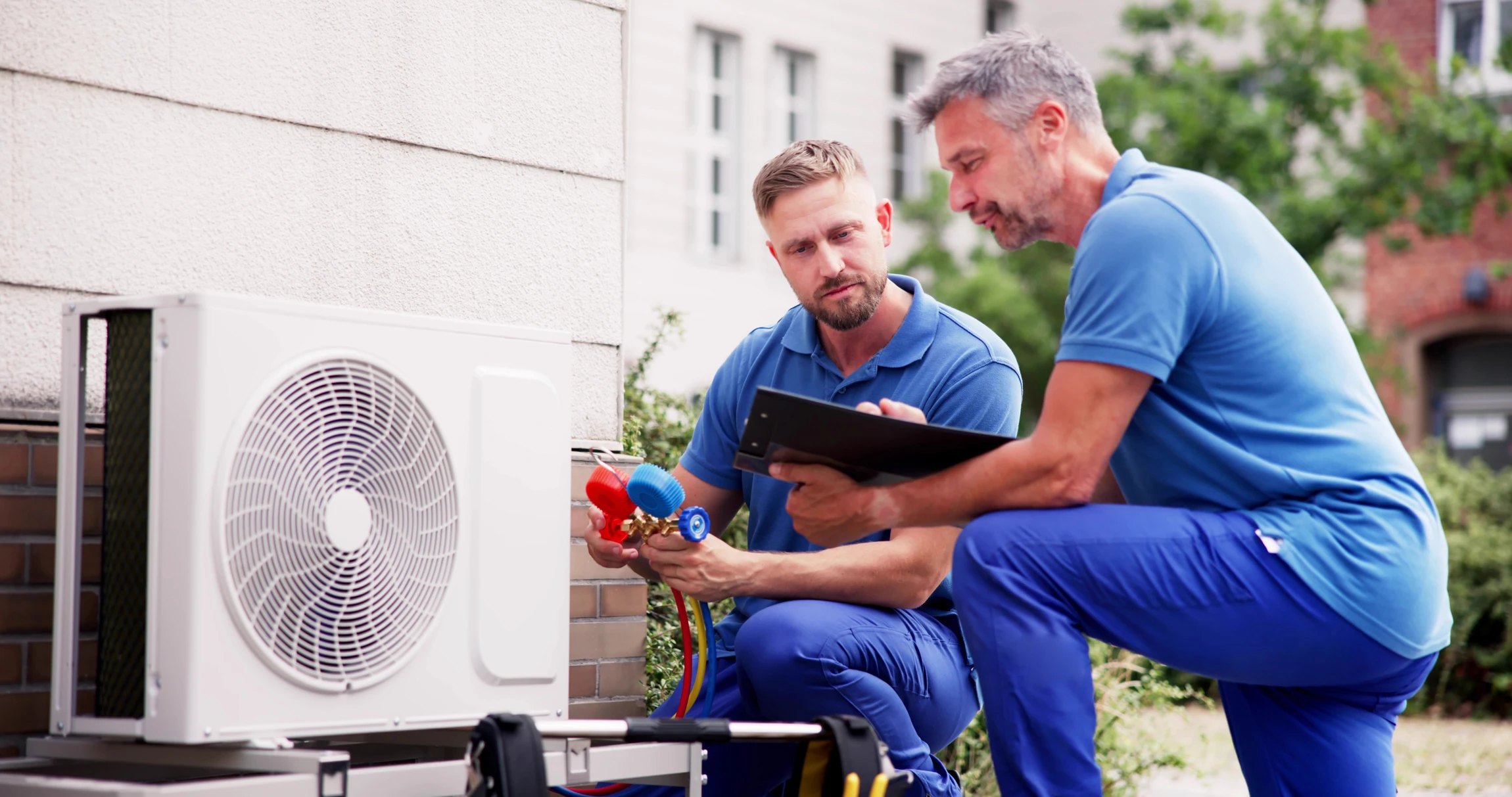 Deux techniciens en uniforme bleu travaillent sur une unité extérieure de climatisation dans un environnement urbain