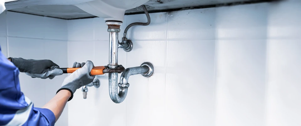 Intérieur sous lavabo, plombier en gants gris utilisant une clé à molette orange sur siphon chromé