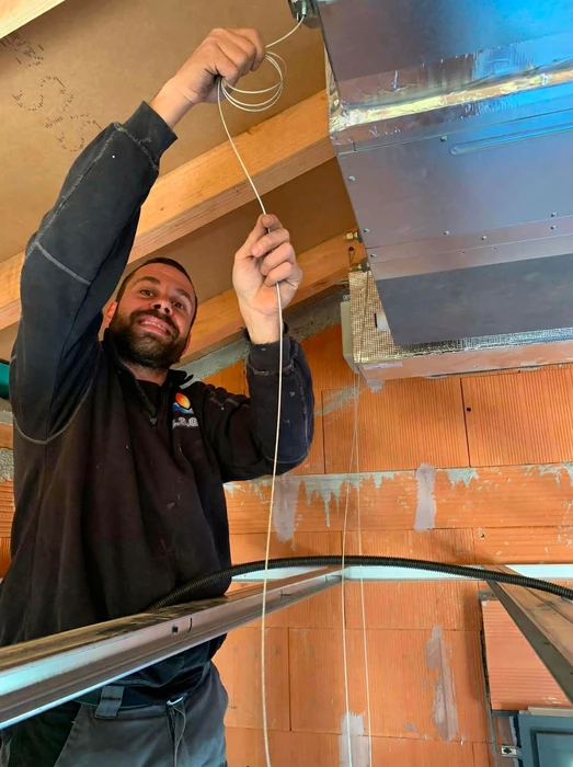 Technicien souriant en combinaison noire, debout sur échafaudage, tirant un câble de signal blanc vers un caisson de ventilation métallique fixé au plafond