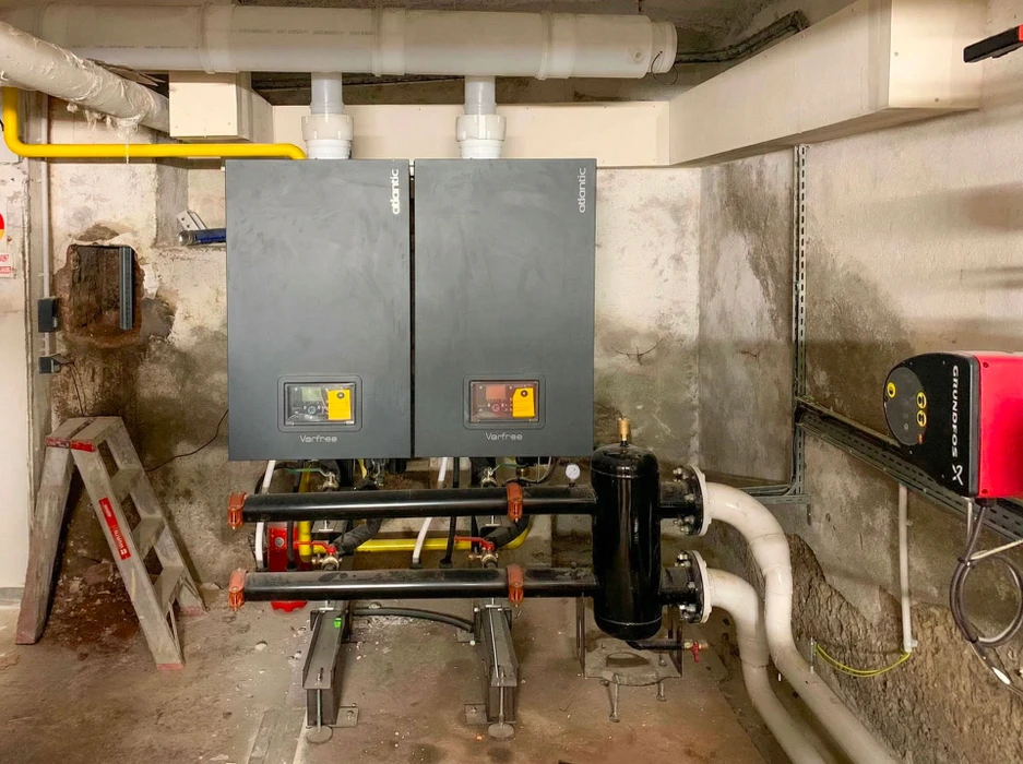 Salle technique intérieure abritant une chaudière biomasse industrielle **KSM Stoker** rouge et grise, avec convoyeur à vis, cyclone de dépoussiérage noir, ventilateur centrifuge bleu, vases d'expansion bleus, réseau de tuyauteries cuivre et acier